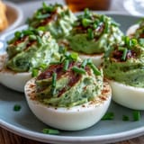 Avocado deviled eggs with smooth, green filling, served on a white platter with a sprinkle of paprika and fresh herbs.  