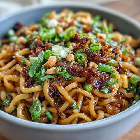 A bowl of chewy wheat noodles coated in spicy chili crisp sauce, garnished with scallions and roasted peanuts for crunch.