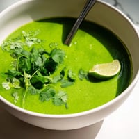 Vibrant spinach coriander lemongrass soup in a white bowl, garnished with fresh cilantro and lime wedges.