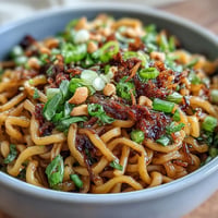 A bowl of chewy wheat noodles coated in spicy chili crisp sauce, garnished with scallions and roasted peanuts for crunch.